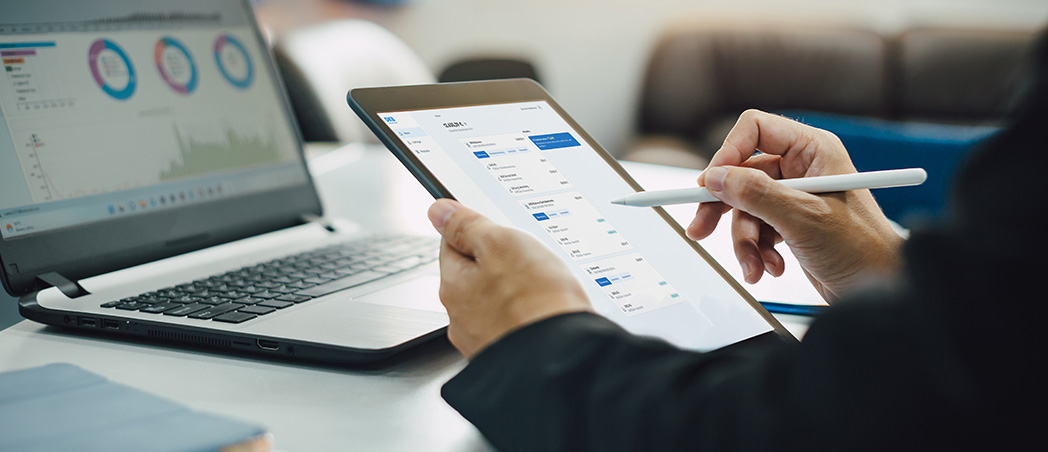 Person analysiert Finanzdaten auf einem Tablet neben einem Laptop im Büro